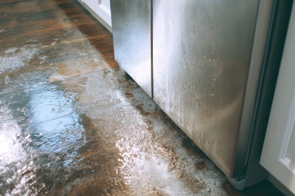 Water leaking from a stainless steel refrigerator onto a wet wooden kitchen floor, causing potential water damage